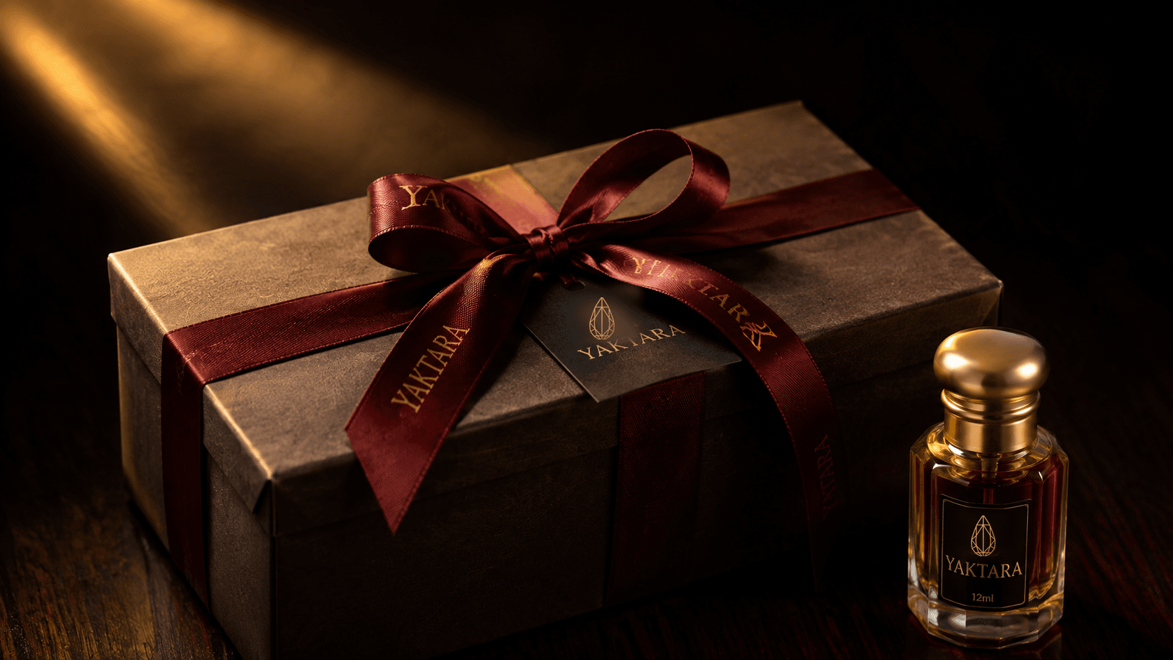 A wrapped Yaktara gift box with a crimson ribbon and branded tag beside a 12ml attar bottle, lit by a warm golden light.