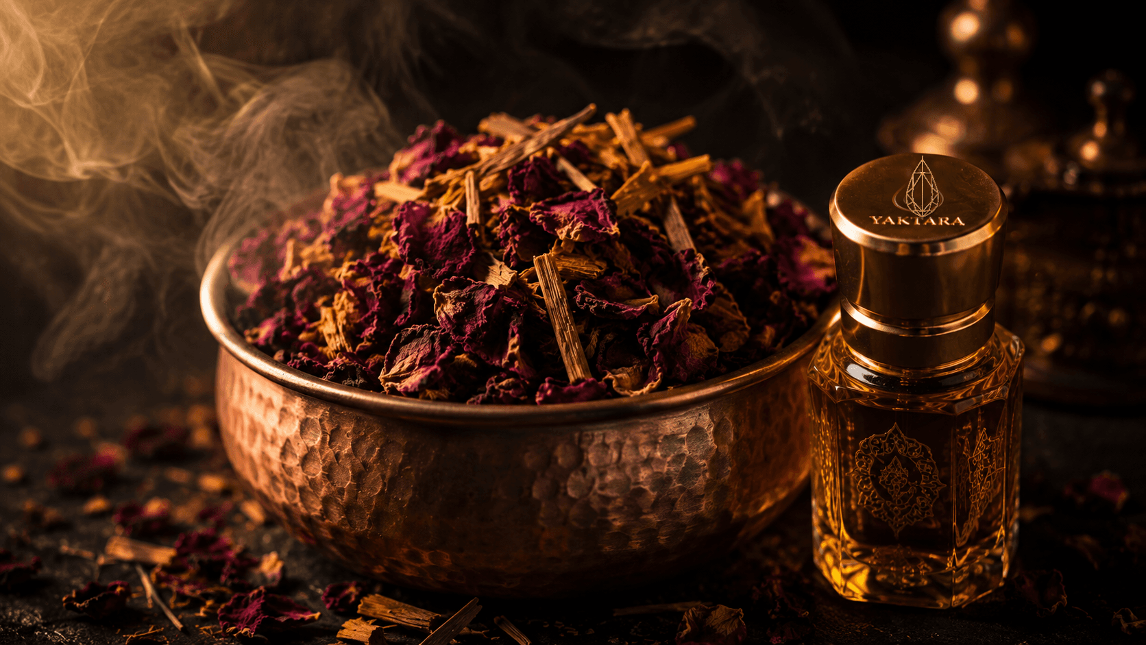 Dried rose petals and sandalwood shavings in a hammered copper bowl beside a Yaktara attar bottle with the drop-mark logo engraved on its gold cap, steam rising in warm light.